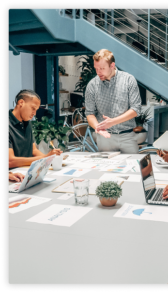 Two people collaborating at a table covered with paperwork, with laptops and other hands visible in the workspace.