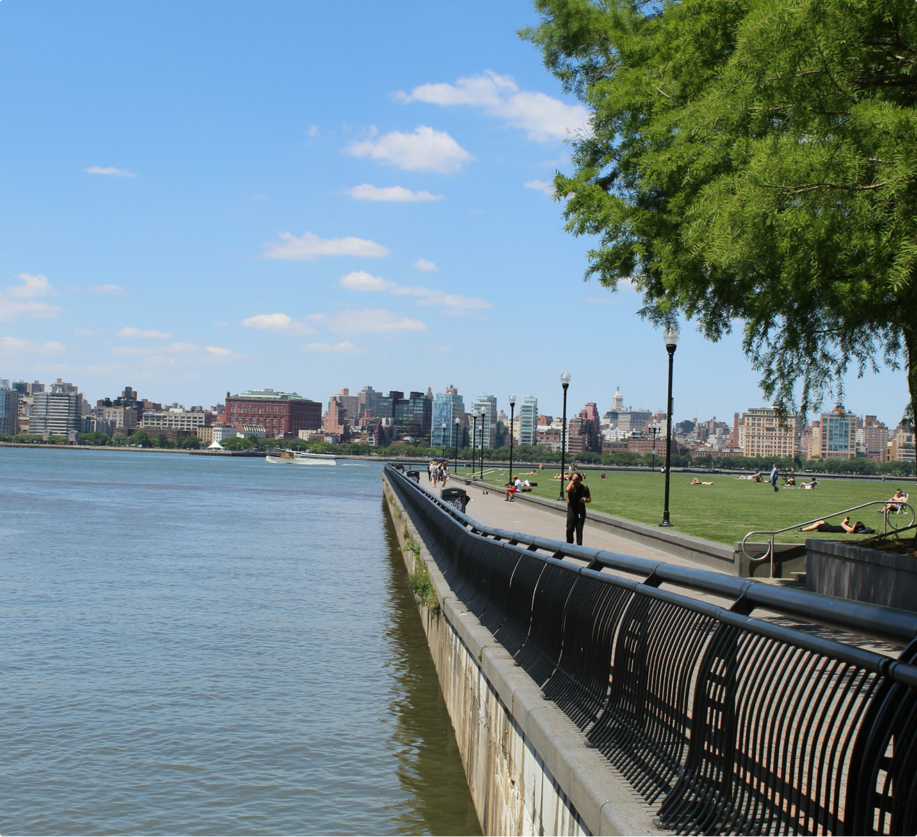 Scenic view along the Hoboken waterfront walkway with railings, green park space, and views of the city skyline across the Hudson River.