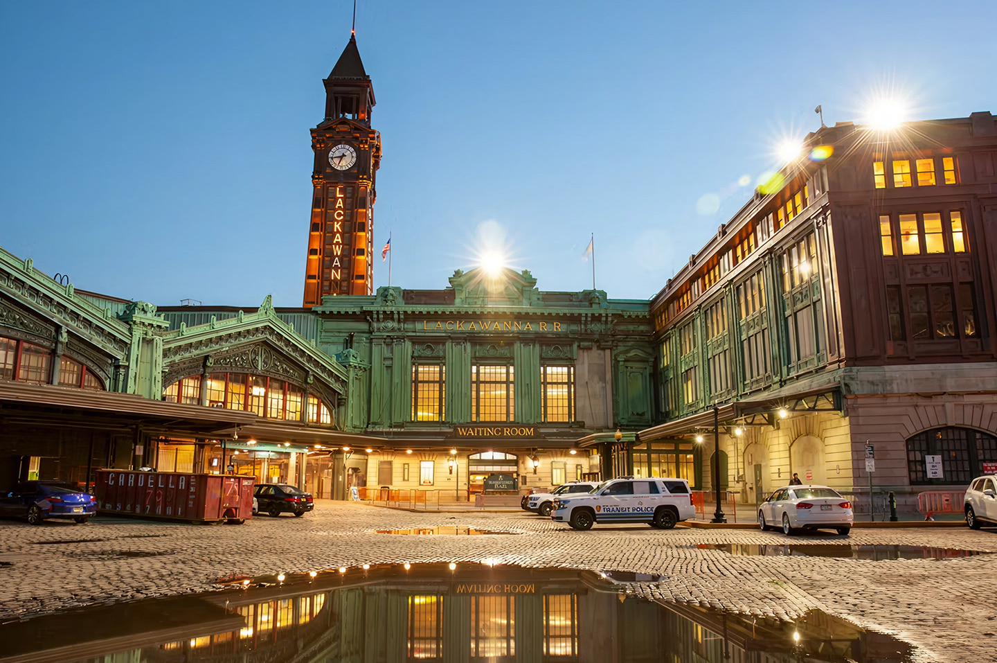 Historic Hoboken Terminal with clock tower and green copper façade illuminated at dawn, with reflections in the wet cobblestone plaza.