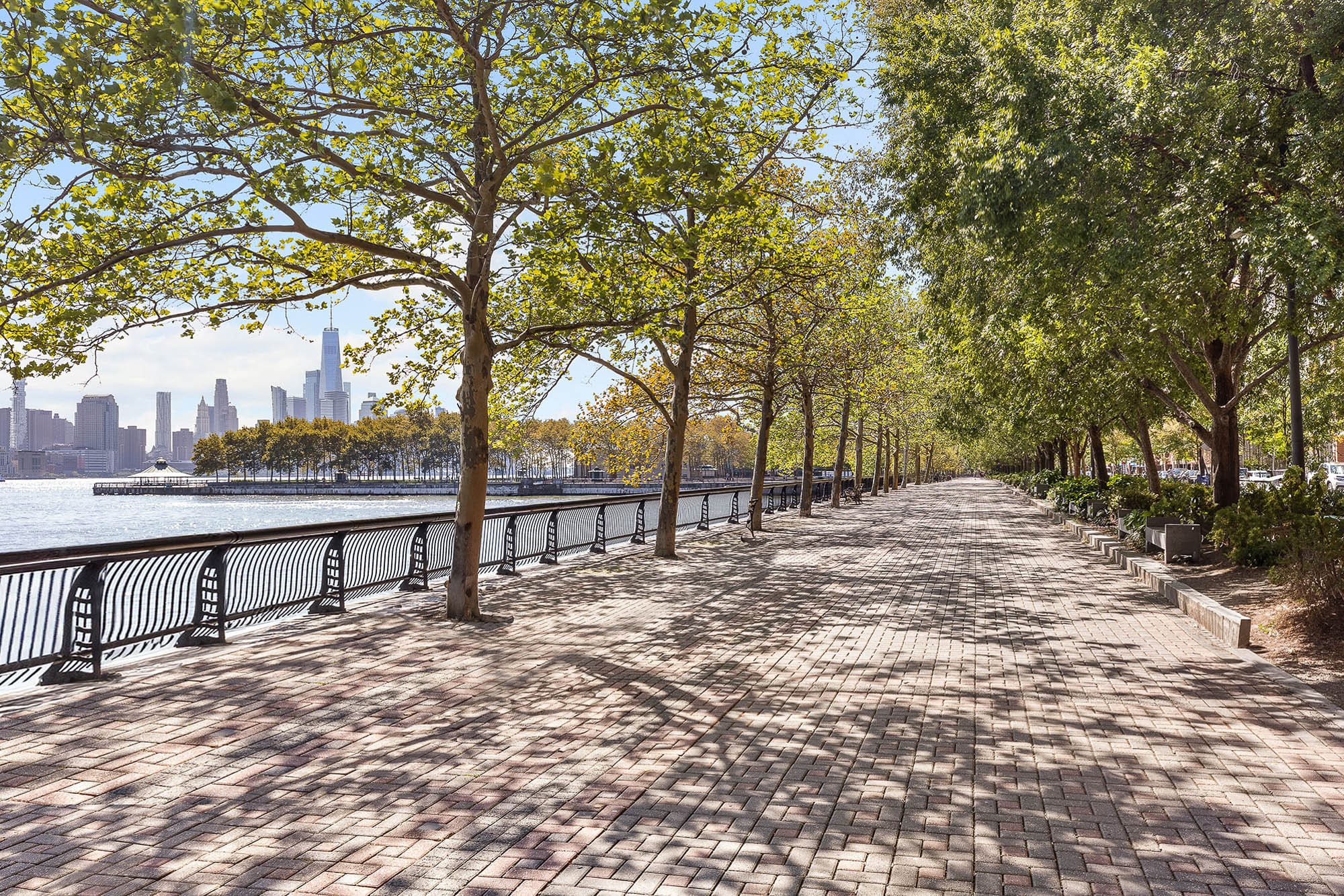 Tree-lined brick promenade along the Hoboken waterfront with views of the Hudson River and Manhattan skyline through the leaves.